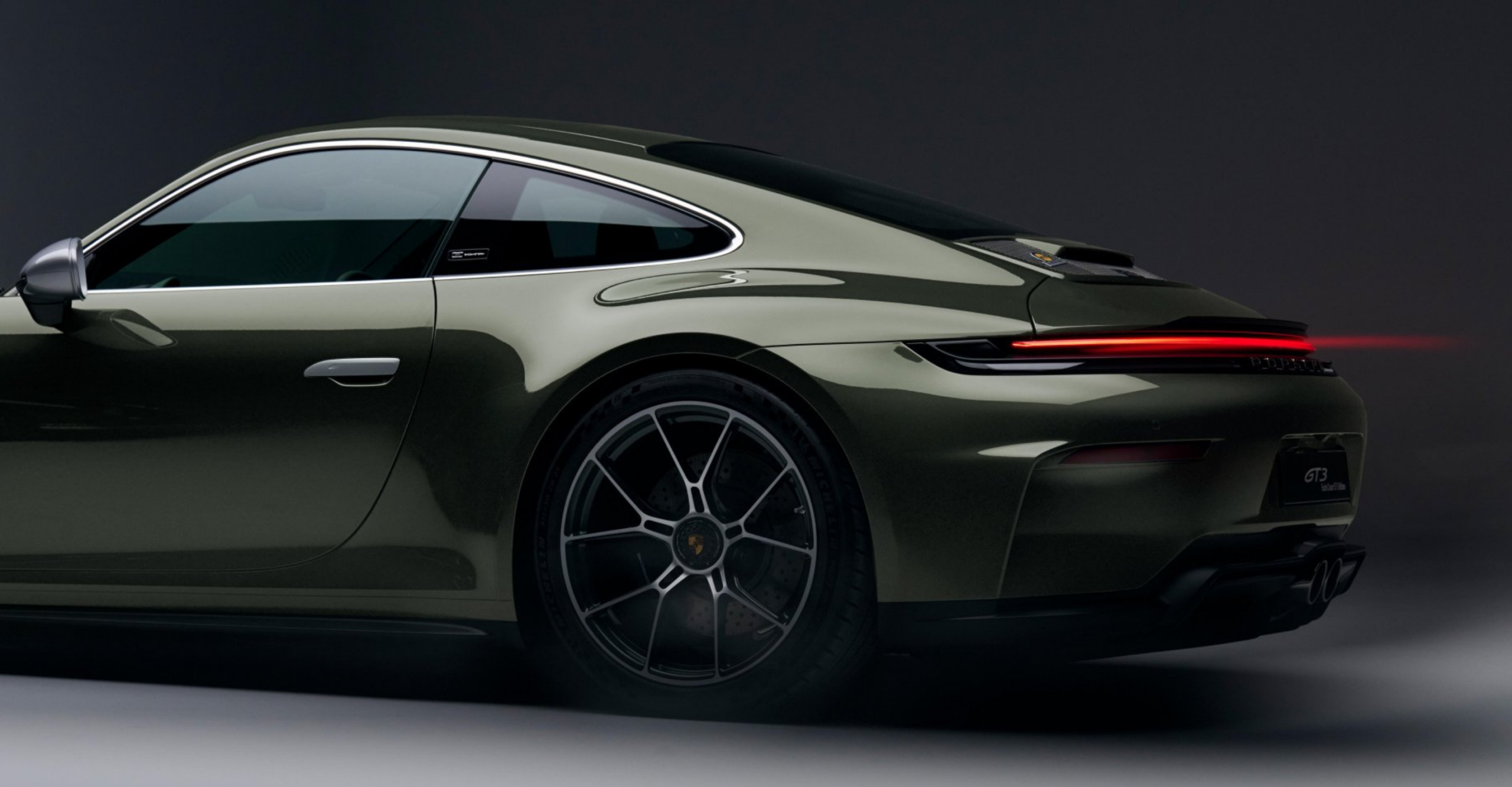 Side profile of an olive green Porsche 911 GT3, highlighting the rear light strip and large alloy wheel on the coupe barrier-free view.